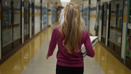 A middle-aged woman with long hair carries books while walking through a school corridor. Bright...