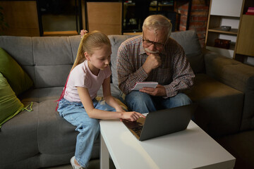 Young girl helping elderly man with laptop while studying together on sofa in cozy home setting....