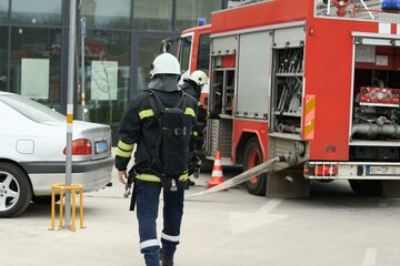 A firefighter in full protective turnout gear walks toward a red fire engine with its compartments open and a hose connected, next to a silver car in a parking lot.