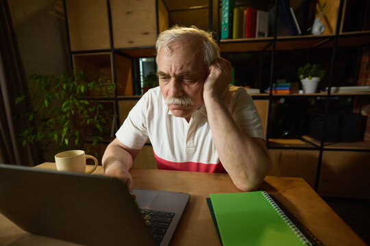 Elderly man with mustache sitting at wooden desk, looking down at laptop with one hand resting on his head, mug and notebooks nearby. Concept of elderly lifestyle, education, knowledge