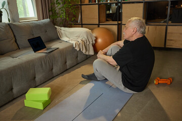Elderly man sitting on yoga mat looking at laptop after completing virtual exercise session at...