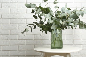 Green eucalyptus branches in vase on table near white brick wall indoors, space for text