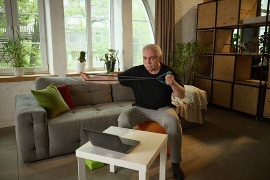 Senior man training arm muscles with resistance band during online workout class at home, training online. Concept of elderly lifestyle, education, knowledge, sport, wellness.