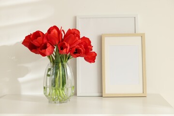 Beautiful red tulips in vase and frames on white table indoors