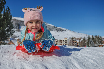 Girl in funny hat sledding on snowy hill under clear blue sky
