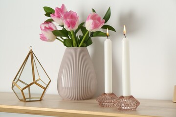 Elegant holders with burning candles, tulips in vase and other decor on wooden table near white wall