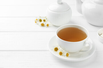 Delicious chamomile tea in cup, fresh flowers and teapot on white wooden table, closeup. Space for text