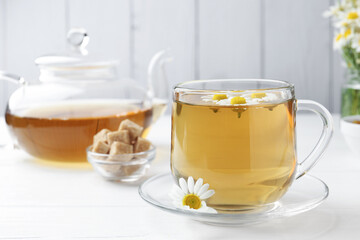 Delicious chamomile tea in glass cup, brown sugar, teapot and fresh flowers on white wooden table, closeup
