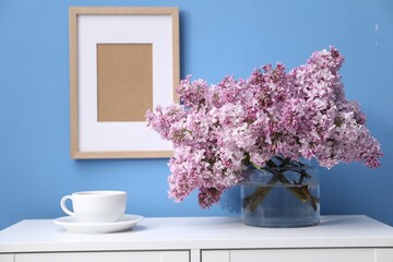 Beautiful lilac flowers in glass vase and cup on cabinet near light blue wall