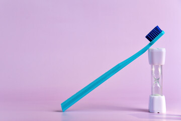 Blue toothbrush leans against a white sand timer on a pastel pink background, illustrating the recommended two-minute oral brushing time