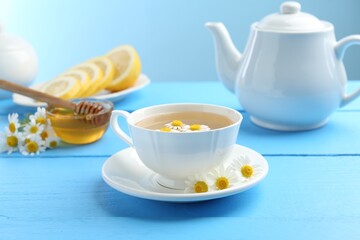 Delicious chamomile tea in cup, fresh flowers, lemon and honey on light blue wooden table, closeup