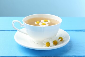 Delicious chamomile tea in cup and fresh flowers on light blue wooden table, closeup