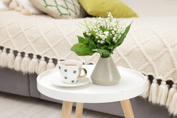 Bouquet of beautiful lily-of-the-valley in vase and cup of tea on coffee table indoors