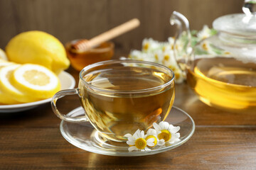 Delicious chamomile tea in glass cup, fresh flowers, lemon and honey on wooden table, closeup