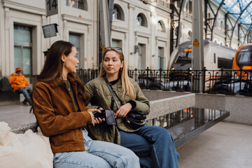 Two women sitting on a bench and talking while holding their phones, at a train station