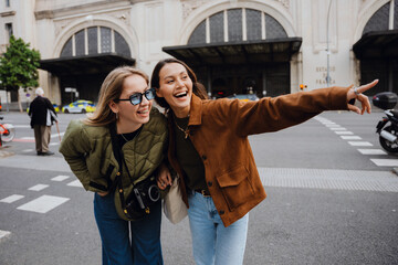 Two female friends stand and laugh while looking in the direction one of them is pointing, on the...