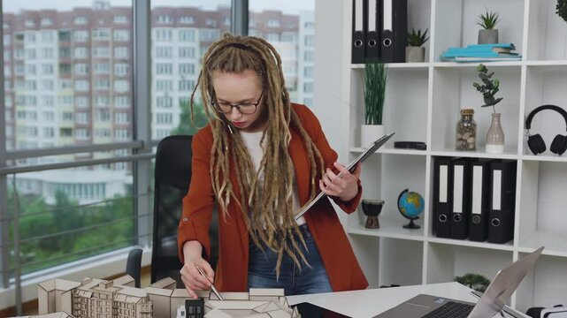 Woman with dreadlocks doing notes into folder about technical construction of new buildings in design studio