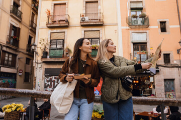 A woman stands and holds street food while looking at a map held by her female friend standing next to her, on the street