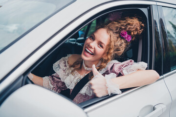 A joyful young woman in an elegant baroque dress gives a thumbs up while sitting in a modern car, blending past and present fashion