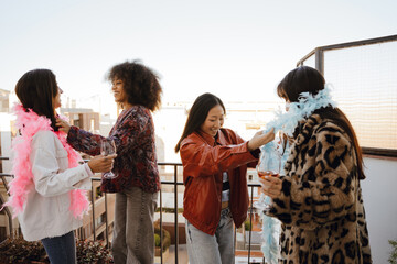A group of four female friends stand and smile while two of them are wearing Hawaiian accessories while holding glasses, on a balcony