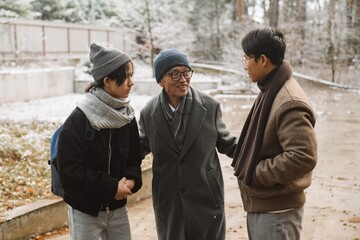 The grandfather is standing and smiling while holding his hands near his son and grandson who are standing next to him, outdoors