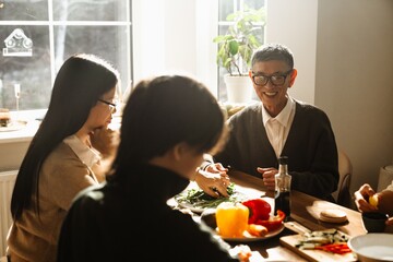 Grandfather sits at the table and smiles while grandmother and grandson sit next to him, they are cooking