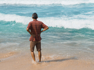 An elderly man stands at the ocean’s edge, hands on hips, gazing at the rolling waves. A moment of reflection and solitude framed by a vivid, sunlit seascape