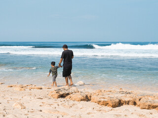 A man and a child hold hands as they walk barefoot along a rocky beach, approaching the gentle...