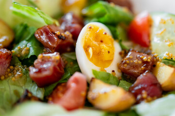 A close-up of a fresh gourmet salad with mixed greens, seared tuna cubes, a halved soft-boiled egg, cherry tomatoes, cucumber slices, and mustard seeds.
