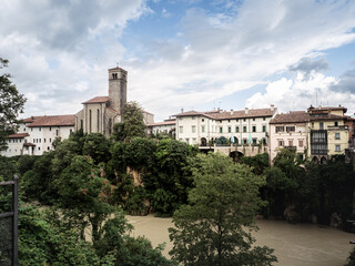 Fototapeta premium Panoramic view of Cividale del Friuli with its church tower and colorful buildings perched above the Natisone River, surrounded by lush greenery and dramatic cliffs.