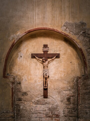 A wooden crucifix depicting Jesus Christ is mounted on an arched, weathered stone wall inside a historic church in Grado, Italy. The worn textures reflect time and reverence.