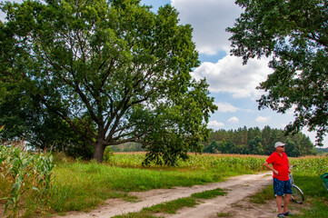 Obraz premium Man farmer standing at village road in countryside with bicycle surrounded with lush green summer nature rural landscape, copy space