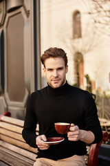 Handsome man enjoying a warm drink while sitting on a bench in a vibrant urban setting