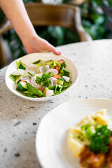 A hand holds a bowl of fresh vegetable salad with radish, cucumber, tomato, and parsley on a speckled countertop. Another plate with mashed potatoes and greens is visible in a bright, inviting setting