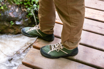 Male legs in trekking boots standing on narrow wooden footbridge over mountain stream during hike