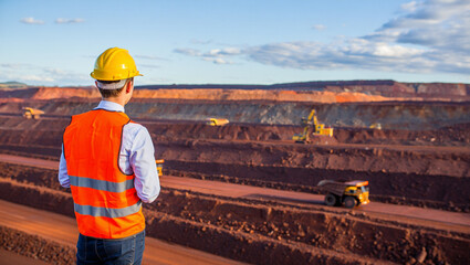 Wide-angle shot of a successful iron ore mining engineer overseeing operations in a massive open pit mine, under clear blue skies, industrial machinery in the background
