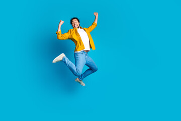 Fototapeta premium Young man joyfully jumping in mid air while expressing excitement on a vibrant blue background wearing casual yellow attire