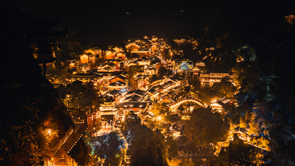 Aerial night view of Wangxian Valley Town, showcasing glowing traditional architecture nestled in the mountains, surrounded by nature and warm ambient lights.