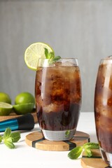 Tasty Cuba Libre cocktail in glasses, ingredients and muddler on white table