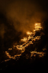 Aerial night view of Wangxian Valley Town, showcasing glowing traditional architecture nestled in the mountains, surrounded by nature and warm ambient lights.