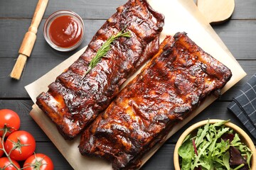 Tasty roasted pork ribs served with rosemary, tomatoes, sauce and salad on black wooden table, flat lay