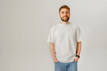 Young man stands casually in a simple white t-shirt with hands in pockets, smiling against a neutral background