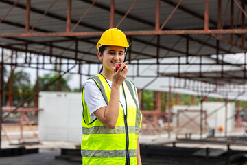 Female civil engineer with walkie talkie stands next to ready made wall or precast boards conducting a quality check with her coworkers in a factory. Engineer in safety suit.