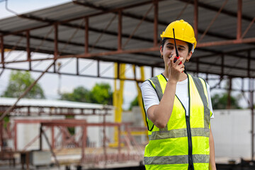 Female civil engineer with walkie talkie stands next to ready made wall or precast boards conducting a quality check with her coworkers in a factory. Engineer in safety suit.