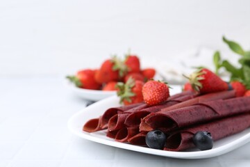Tasty fruit leather rolls, strawberries and blueberries on white table, closeup. Space for text