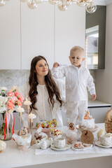 Happy mother and son in white clothes in kitchen with Easter decor. Basket, rabbit, eggs, milk and flowers on table