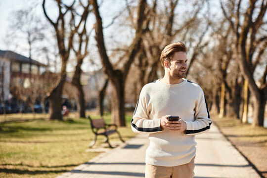 Handsome man walking through a park in warm sunlight while checking his smartphone