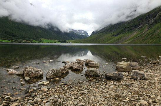 Landscape photo with a view of mountains partially covered in clouds and the mirror-like surface of Eidsvatnet Lake near Geiranger in Norway