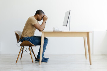 Portrait of stressed African American man sitting at wooden desk with his head in his hands, upset...