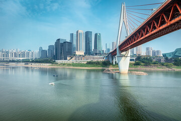 Modern red cable-stayed bridge and contemporary city skyline viewed across the calm river with small boats on a sunny day in Chongqing, China.